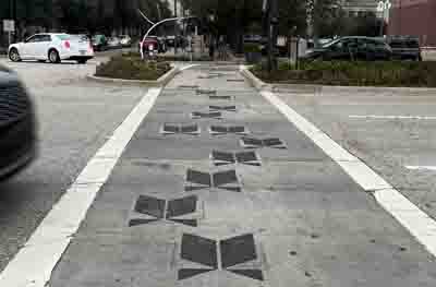 Image is of a crosswalk with 2 thick white lines on the perimeter.  In the center are scattered large black shapes resembling open books, stretching across the entirety of the crossing.